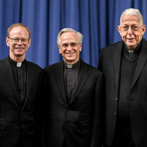 Father Dowd stands with Father Jenkins and Father Malloy. All three are wearing black and standing in front of a blue background