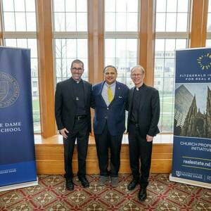 Rev. Robert A. Dowd, C.S.C., stands in front of windows and smiles with two Notre Dame faculty members.