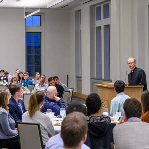 Fr. Dowd stands behind a podium and speaks to a group of students seated at tables