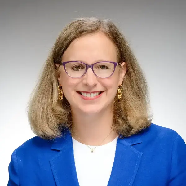 Renée LaReau is a woman with blonde hair and purple glasses. She smiles directly into the camera and wears gold earrings and a blue blazer against a grey background.